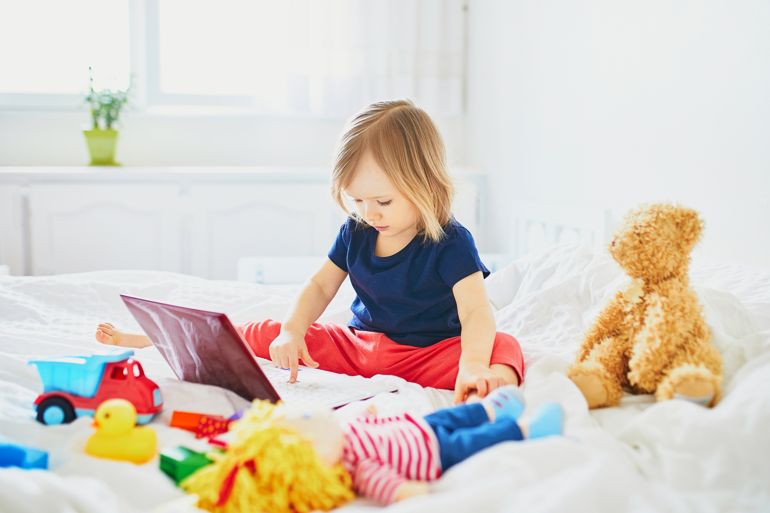 Little Girl Playing with Laptop and Toys on the Bed